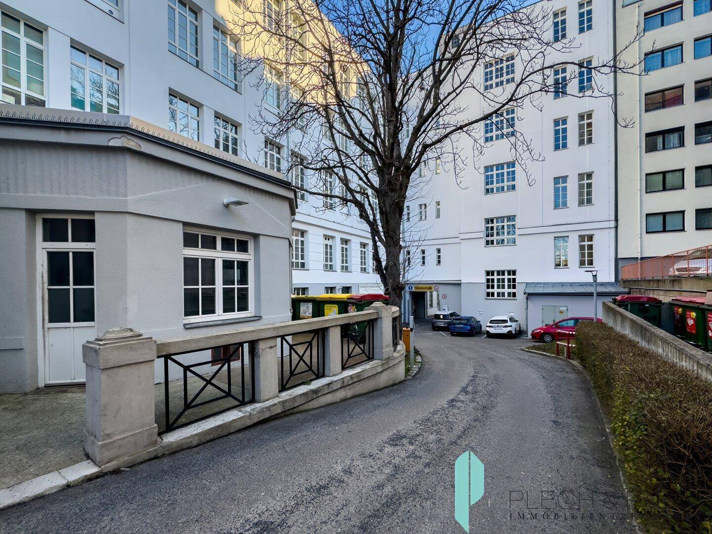 Innenhof mit Zufahrt zu einem Parkplatz, umgeben von der Gebäudefassade und Bäumen.