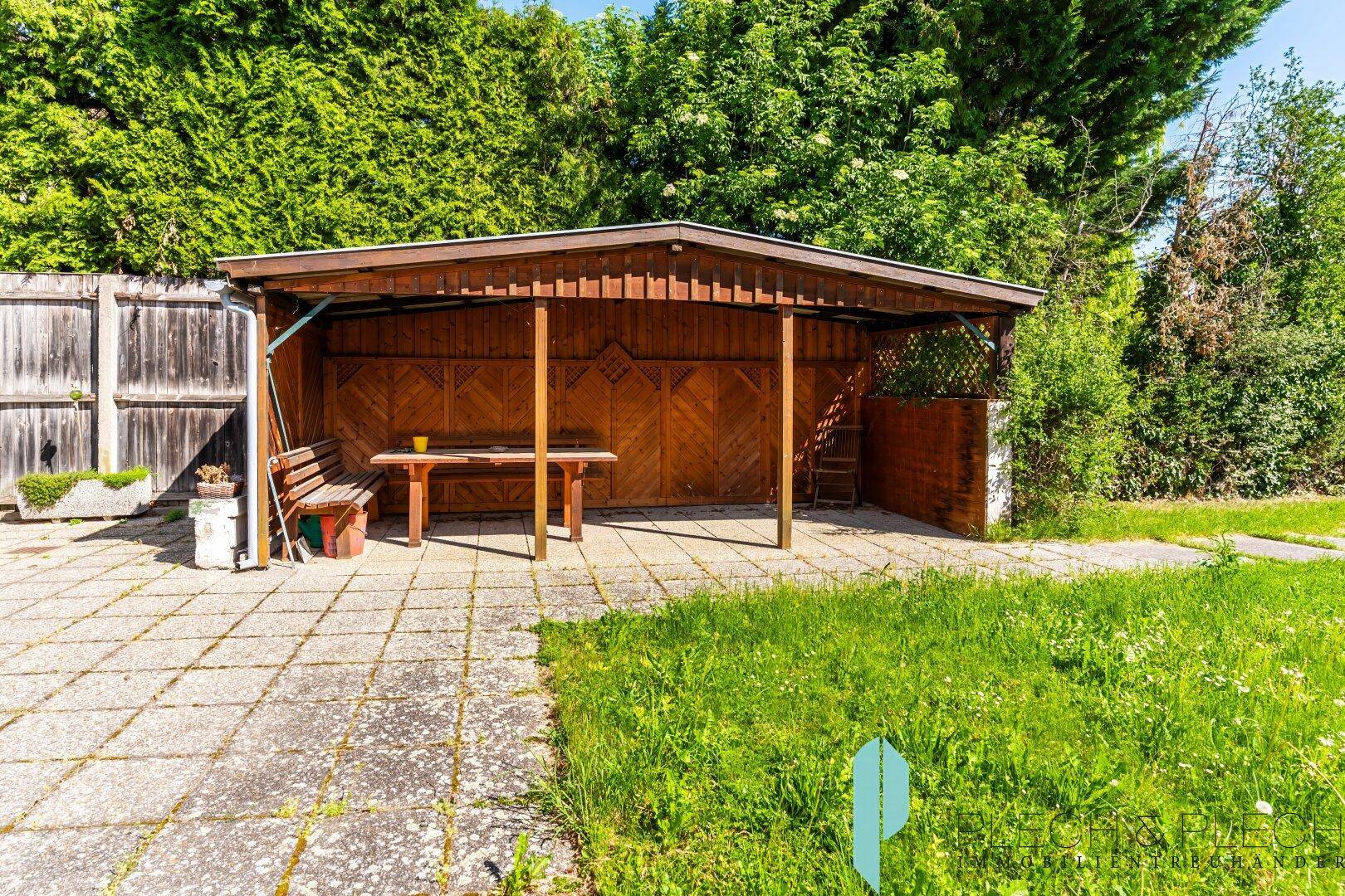 Überdachter Holzunterstand mit Sitzgelegenheiten und gepflastertem Bereich im Garten.