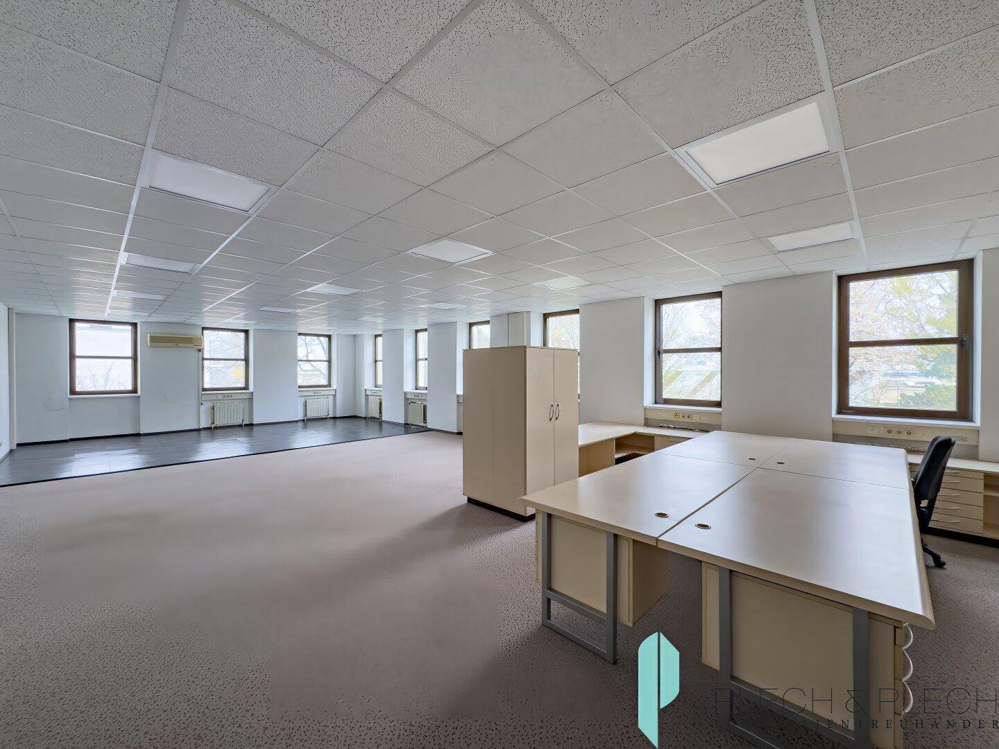 Großer, heller Büroraum mit Teppichboden, abgehängter Decke und mehreren Fenstern, teilweise möbliert.
