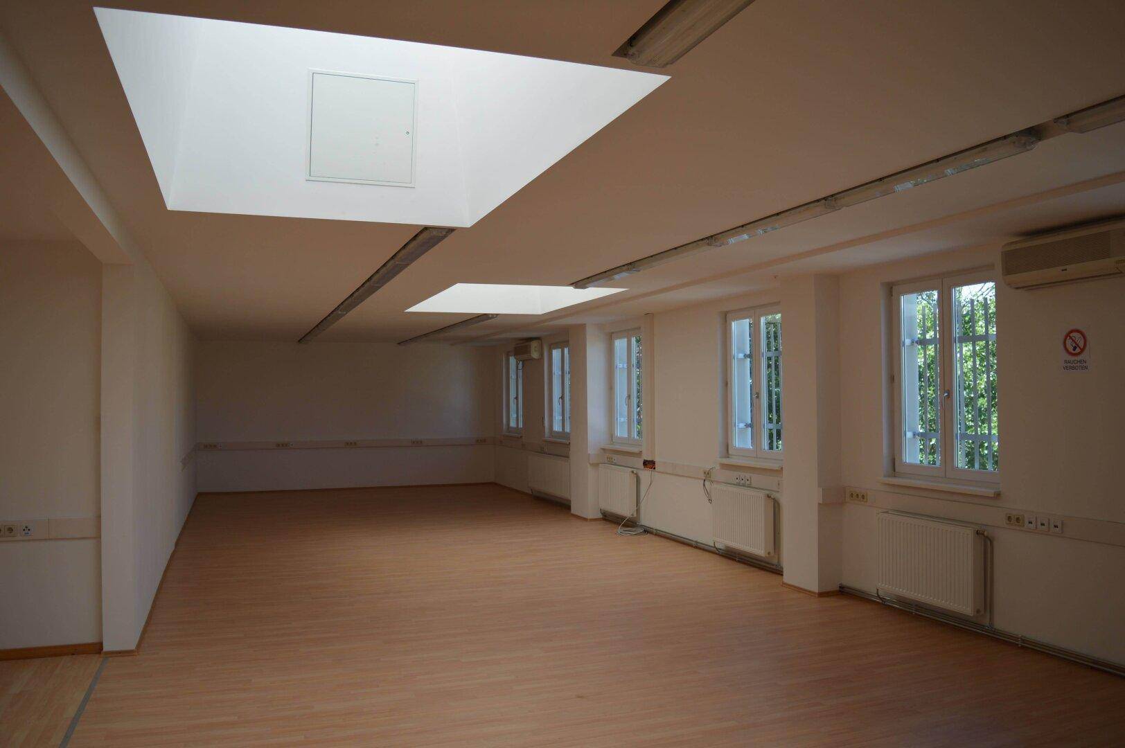 Langer, heller Büroraum mit Holzboden, vielen Fenstern und integrierten Deckenleuchten, sehr geräumig.