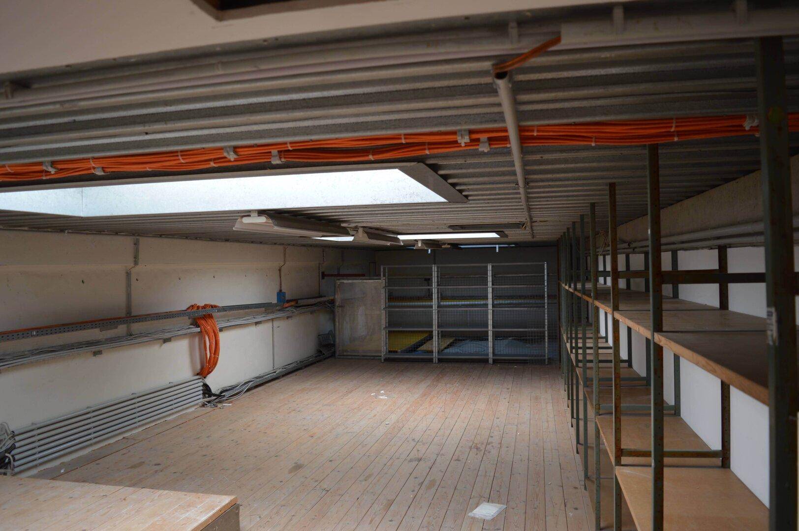 Lagerraum mit Holzfußboden, vielen Regalen und sichtbaren Installationen an der Decke und Wänden.