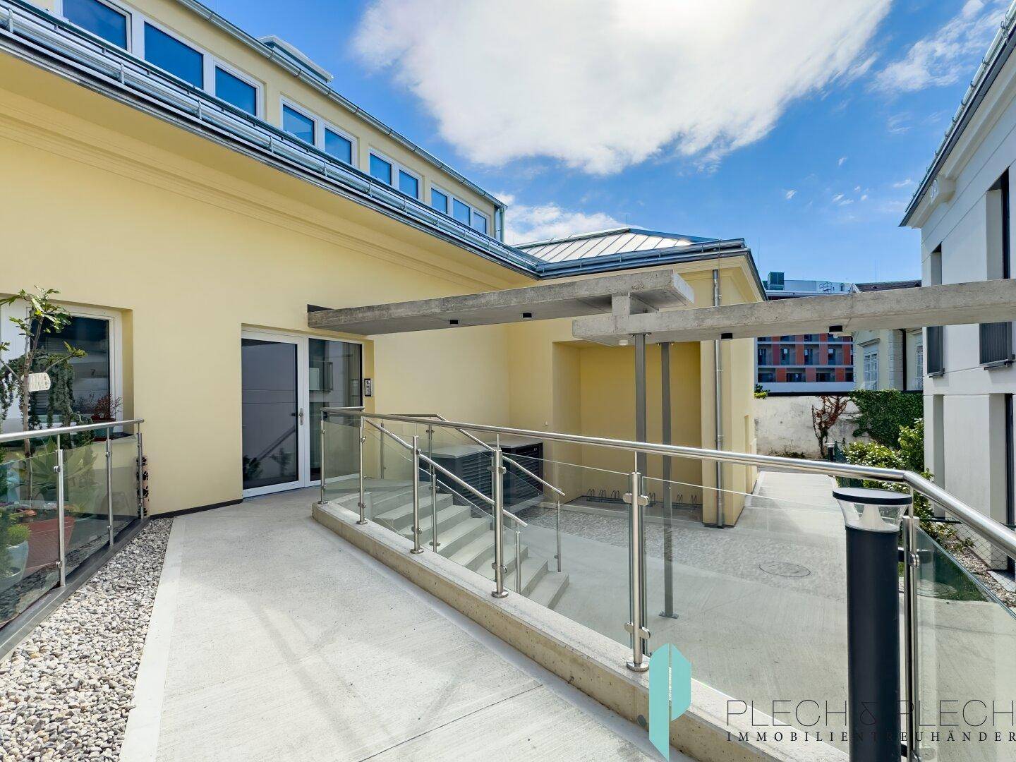 Moderne Terrasse mit Glasgeländer und Treppe, die zu einem Eingang des gelben Gebäudes führt.