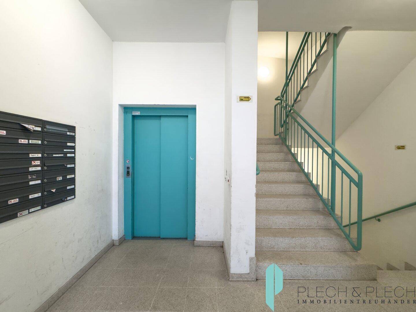 Eingangsbereich des Gebäudes mit Treppenhaus, modernem Aufzug und Briefkästen.
