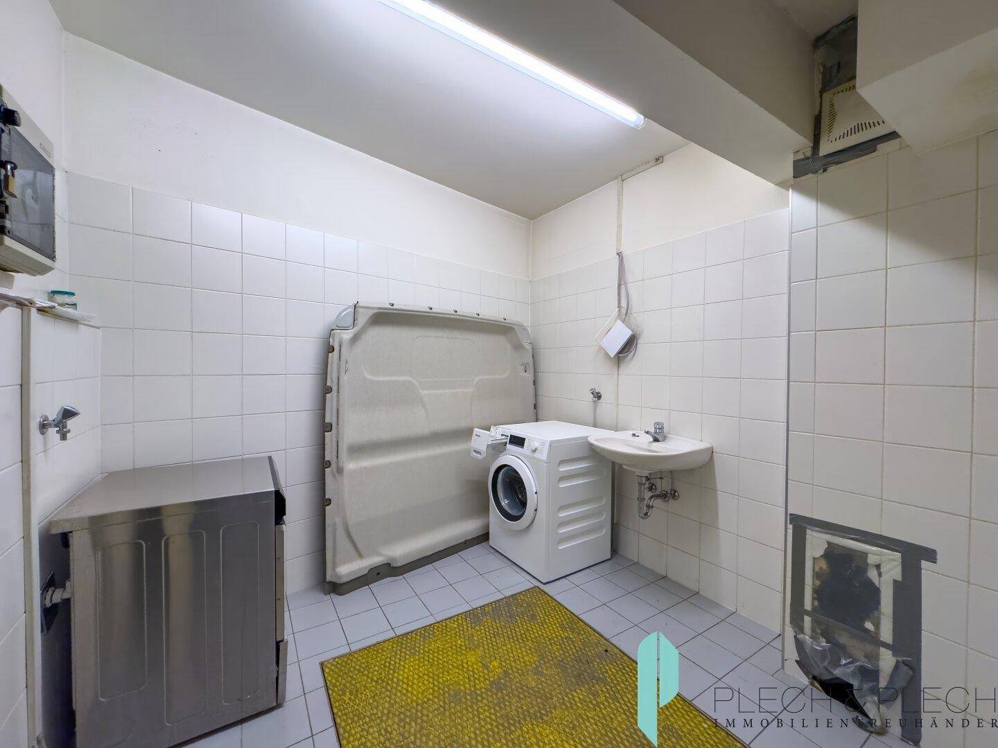 Gemeinschaftlicher Waschraum mit Waschmaschinen und Waschbecken, komplett gefliest für einfache Reinigung.