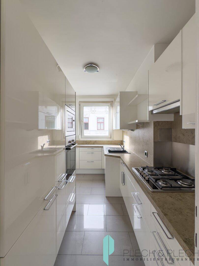 Moderne, schmale Küche mit weißen Hochglanzschränken, Gasherd und Fenster.