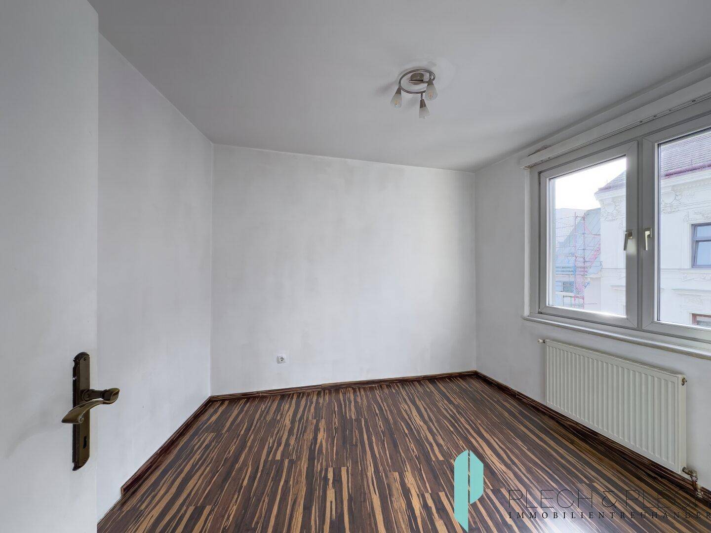 Leeres Schlafzimmer mit dunklem Laminatboden und großem Fenster, das viel Licht hereinlässt.