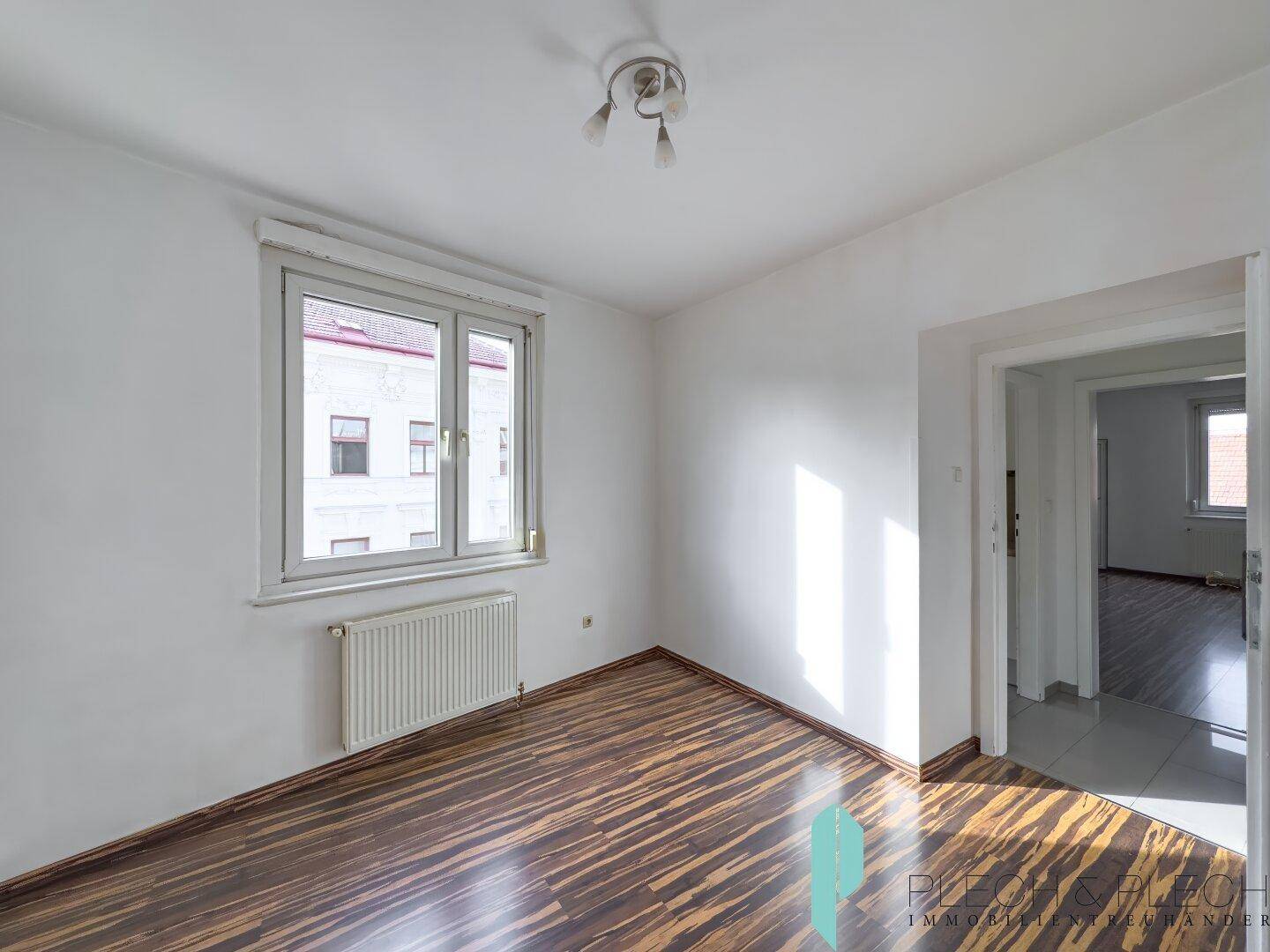 Helles Schlafzimmer mit dunklem Laminatboden und Blick in einen angrenzenden Raum.
