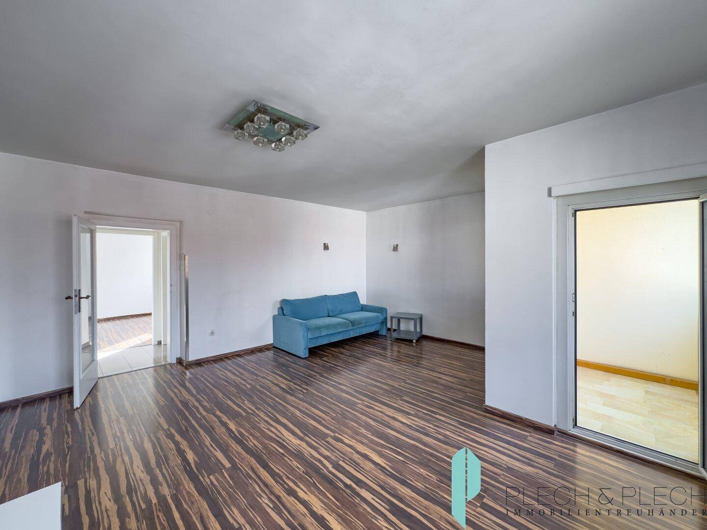 Wohnzimmer mit dunklem Laminatboden, blauer Couch und Blick in angrenzende Räume.