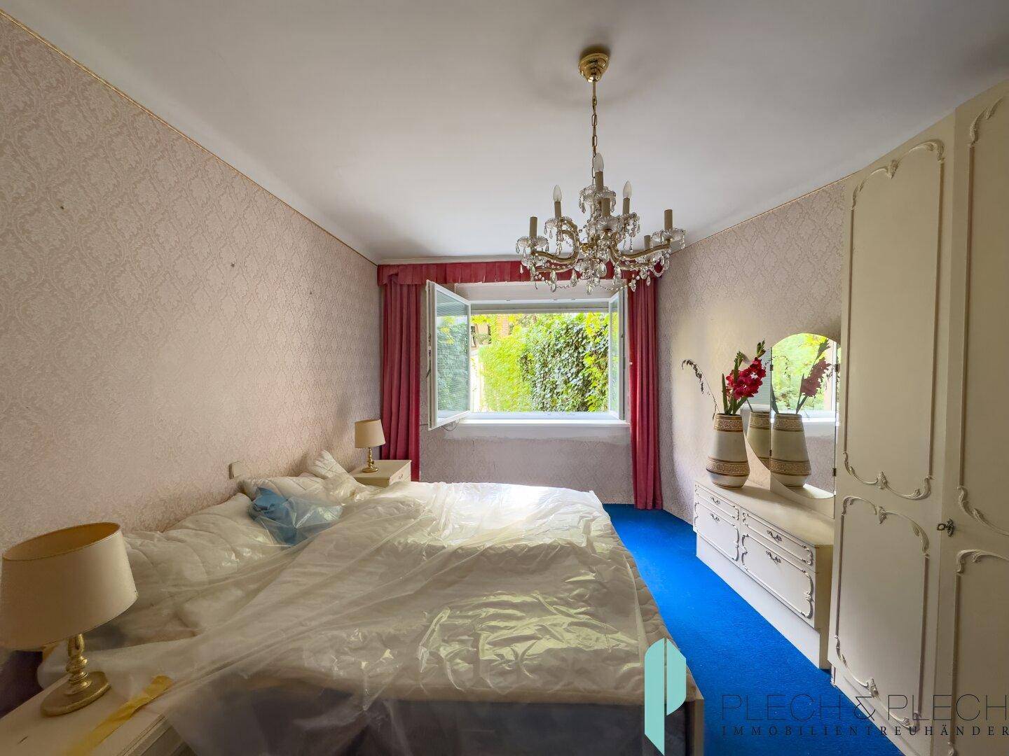 Schlafzimmer mit blauem Teppichboden, Kronleuchter und Blick ins Grüne.