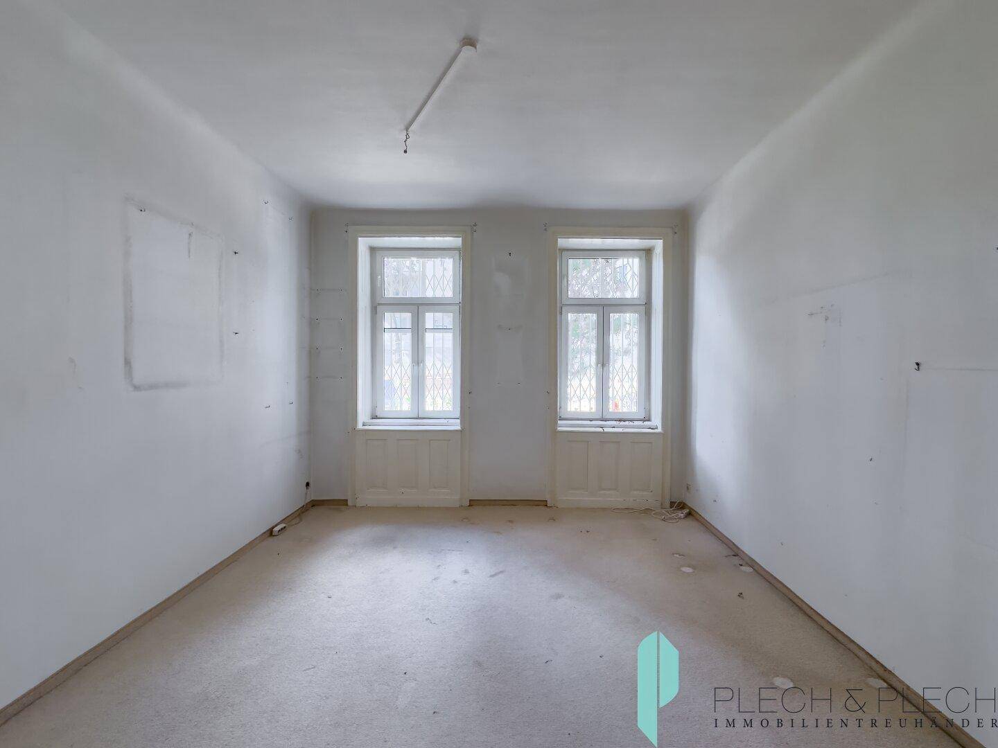 Leeres Zimmer mit Teppichboden und zwei hohen Fenstern, die den Raum erhellen.