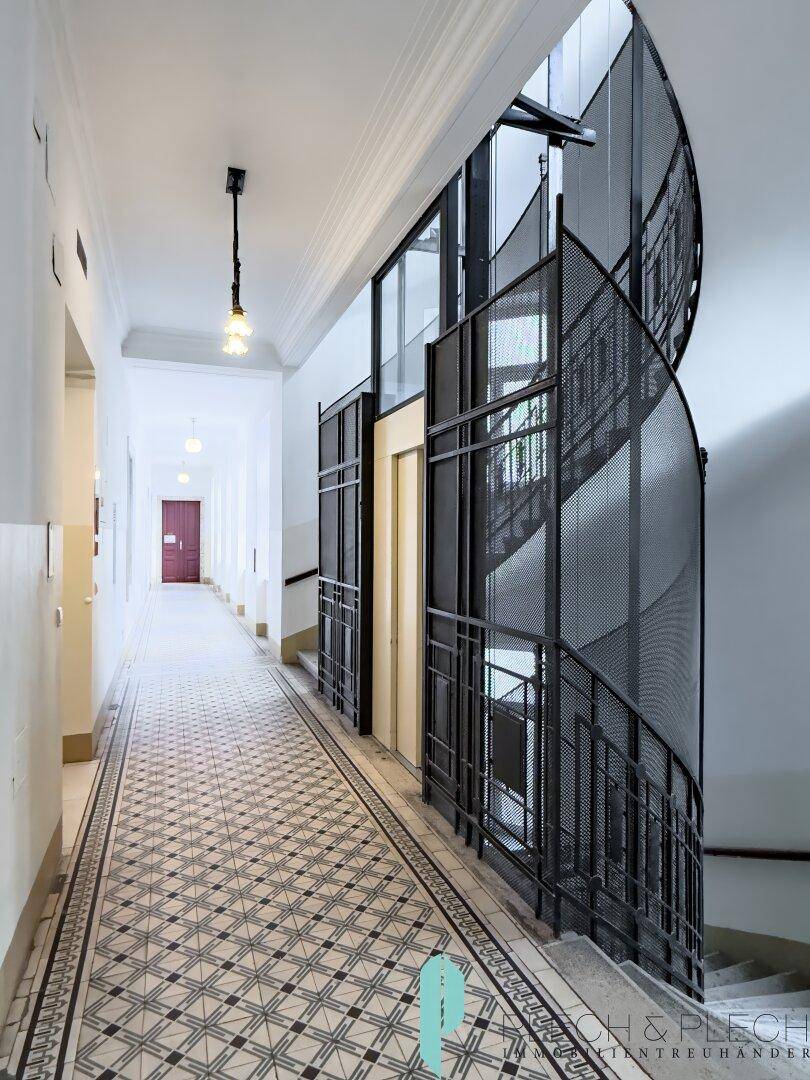Langer Flur mit gemusterten Fliesen, modernem Aufzug und Treppenhaus mit Metallgeländer.
