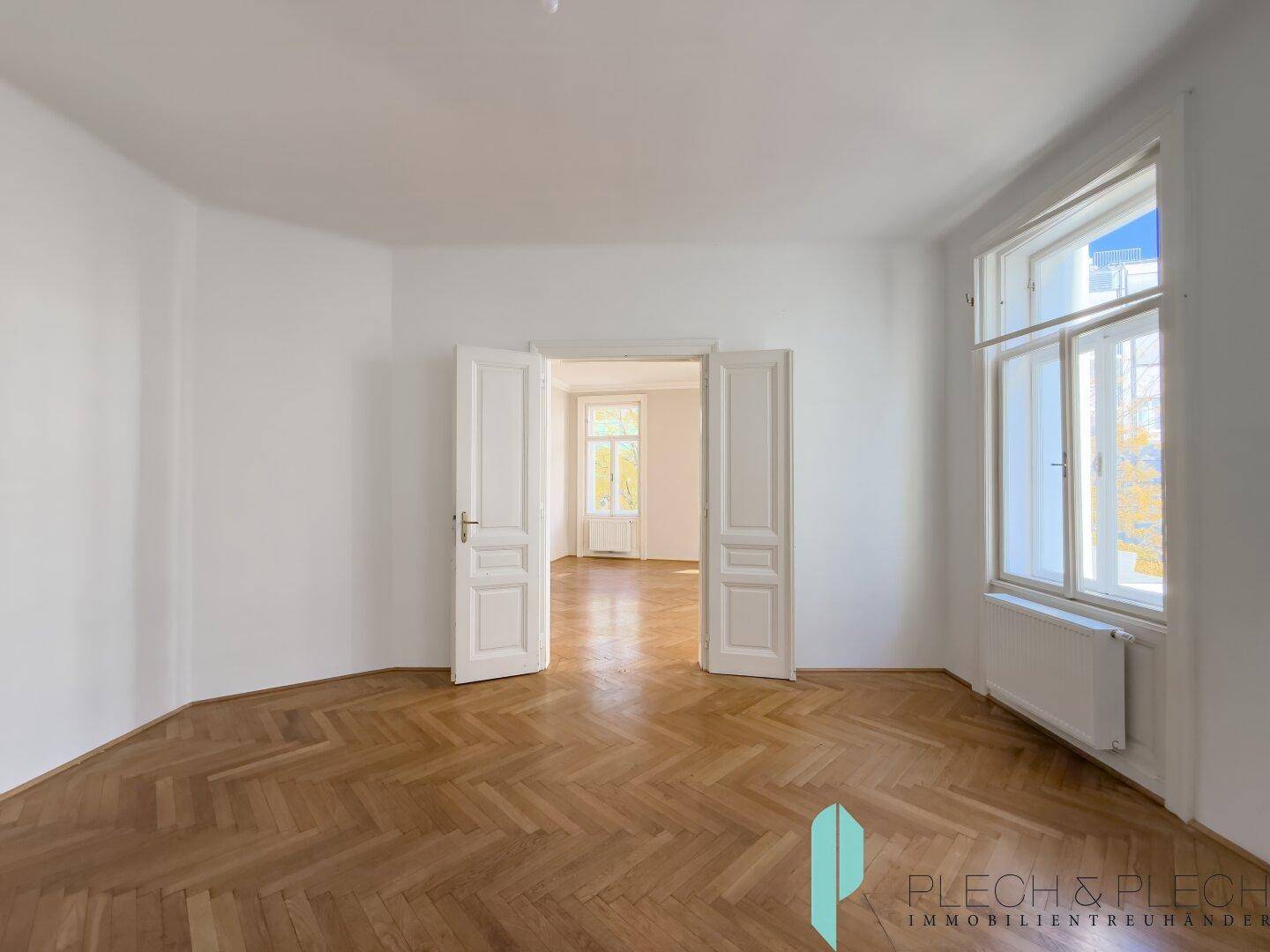 Geräumiger Raum mit Fischgrätparkett und hohen Doppeltüren, die zu einem weiteren Zimmer führen.