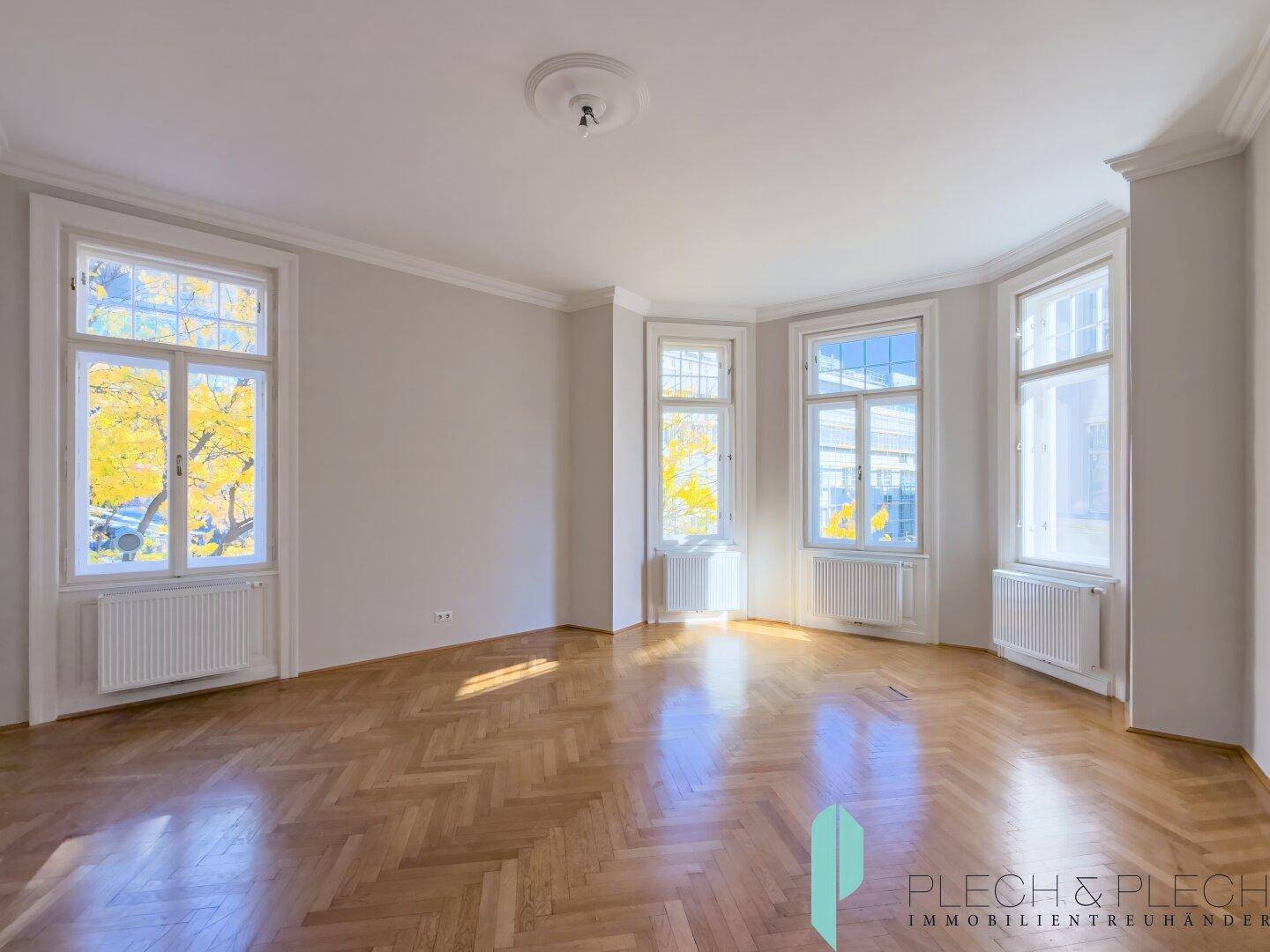 Heller Raum mit Fischgrätparkett, hohen Fenstern und Stuckelementen an der Decke.