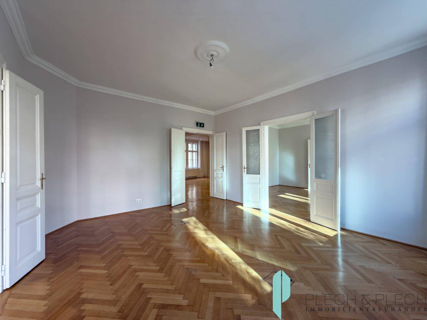 Großer Raum mit Fischgrätparkett, hohen Decken und mehreren Türen, die viel Licht hereinlassen.