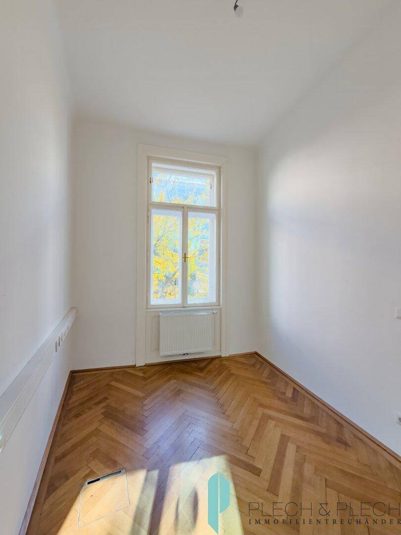 Kleiner, heller Raum mit Fischgrätparkett und einem hohen Fenster, das viel Tageslicht bietet.