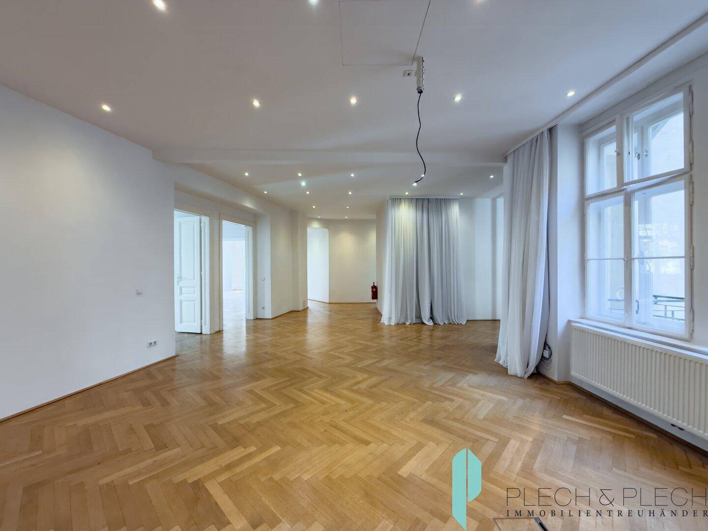 Weitläufiger Raum mit Fischgrätparkett, vielen Einbaustrahlern und Blick auf weitere Zimmer.
