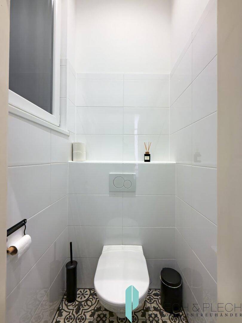 Modernes Gäste-WC mit weißen Fliesen, Wand-WC und gemustertem Boden.