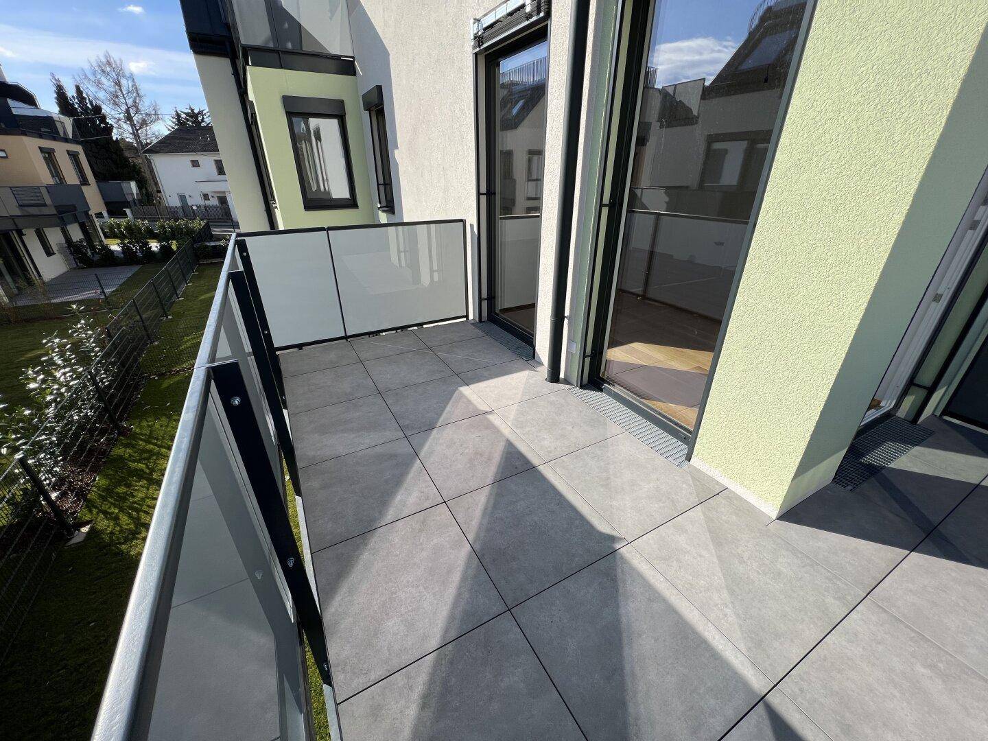 Moderner Balkon mit grauen Fliesen und Glasbrüstung, bietet Platz für Sitzgelegenheiten.