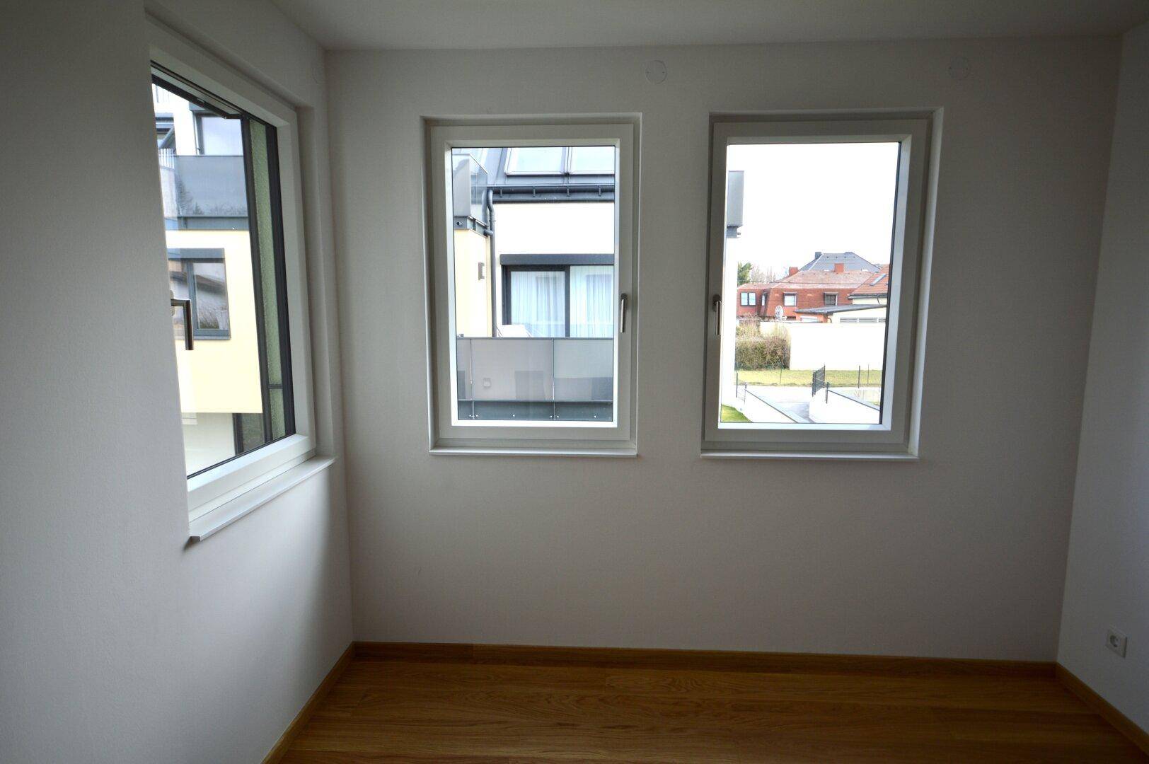 Helles Zimmer mit Holzboden und drei Fenstern, die einen Blick auf die Nachbarschaft bieten.