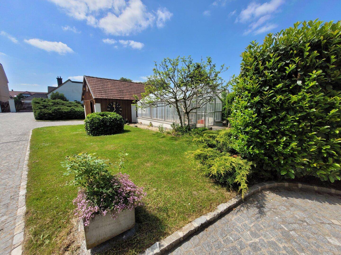 Gepflegter Gartenbereich mit Rasenfläche, Gewächshaus und einem kleinen Schuppen.