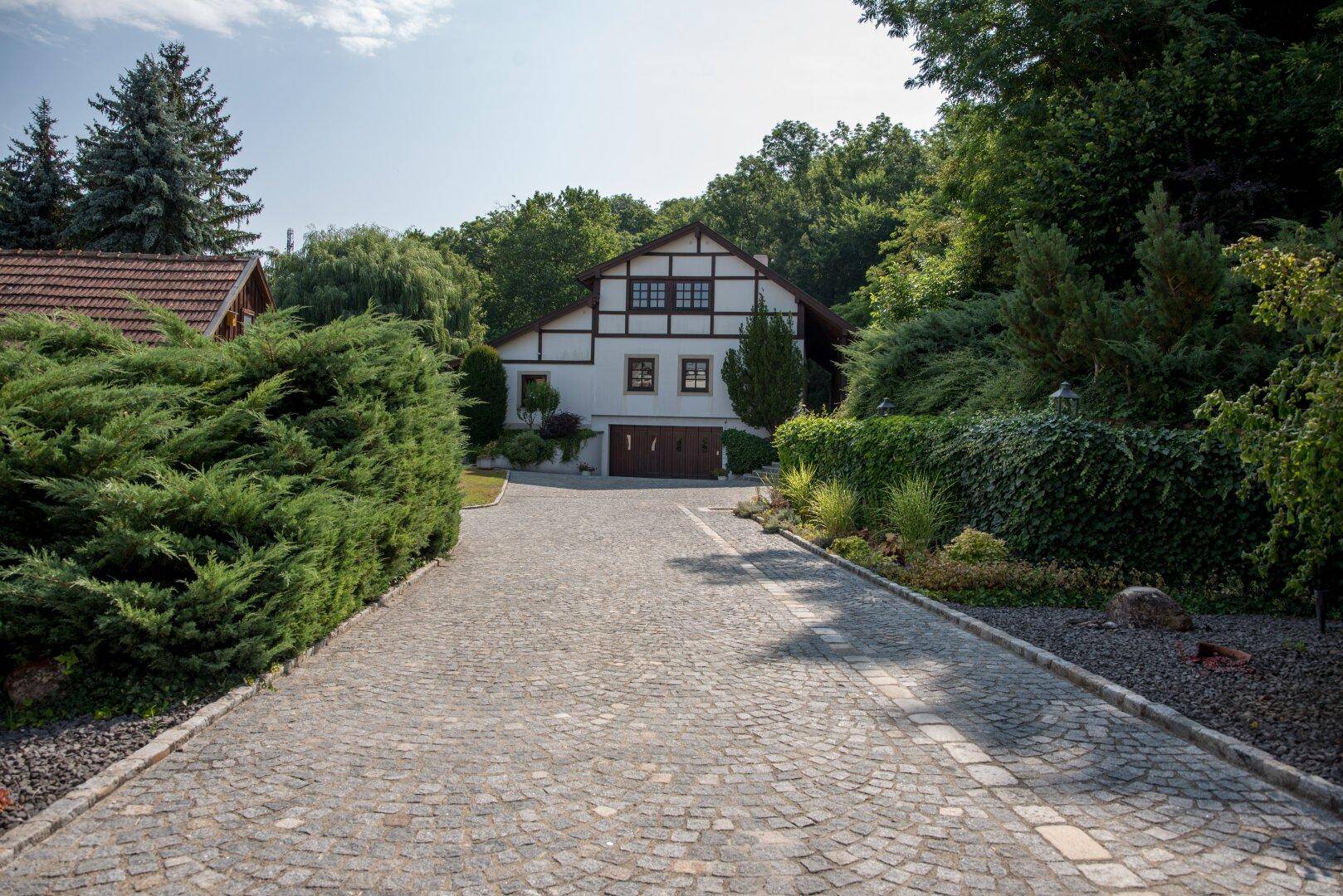 Lange, gepflasterte Auffahrt führt zum Haus, umgeben von Hecken und Bäumen.