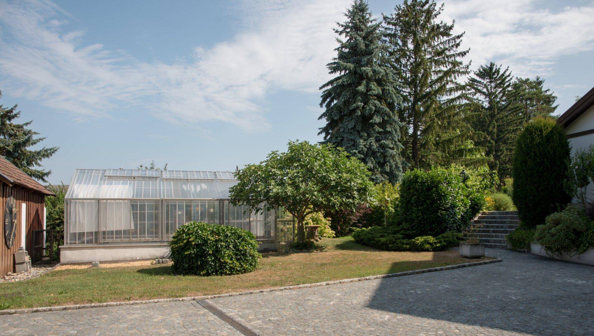 Großes Gewächshaus im Garten, umgeben von gepflegten Pflanzen und Bäumen.