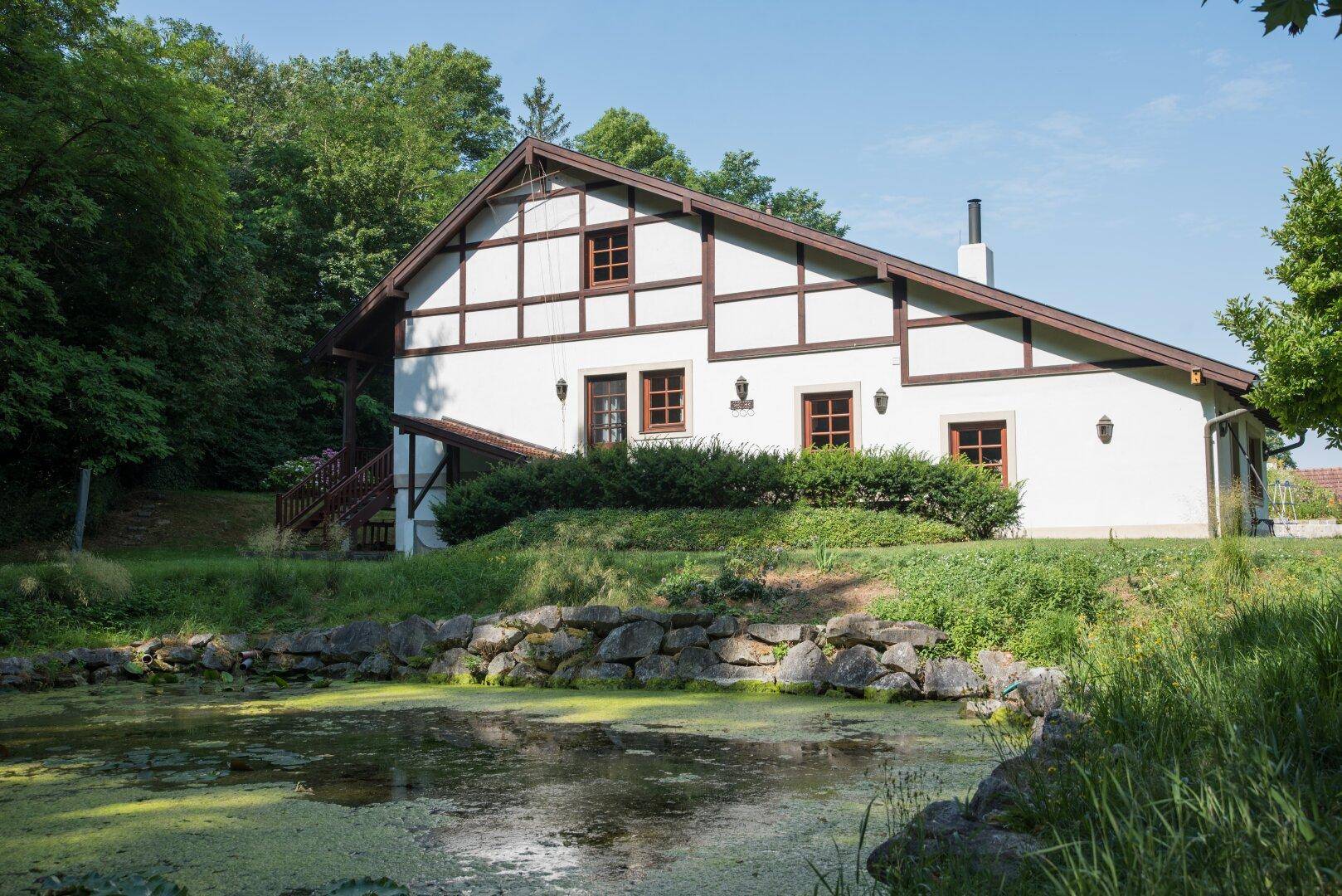 Traditionelles Haus mit Fachwerkfassade und einem Teich im Vordergrund, umgeben von üppigem Grün.