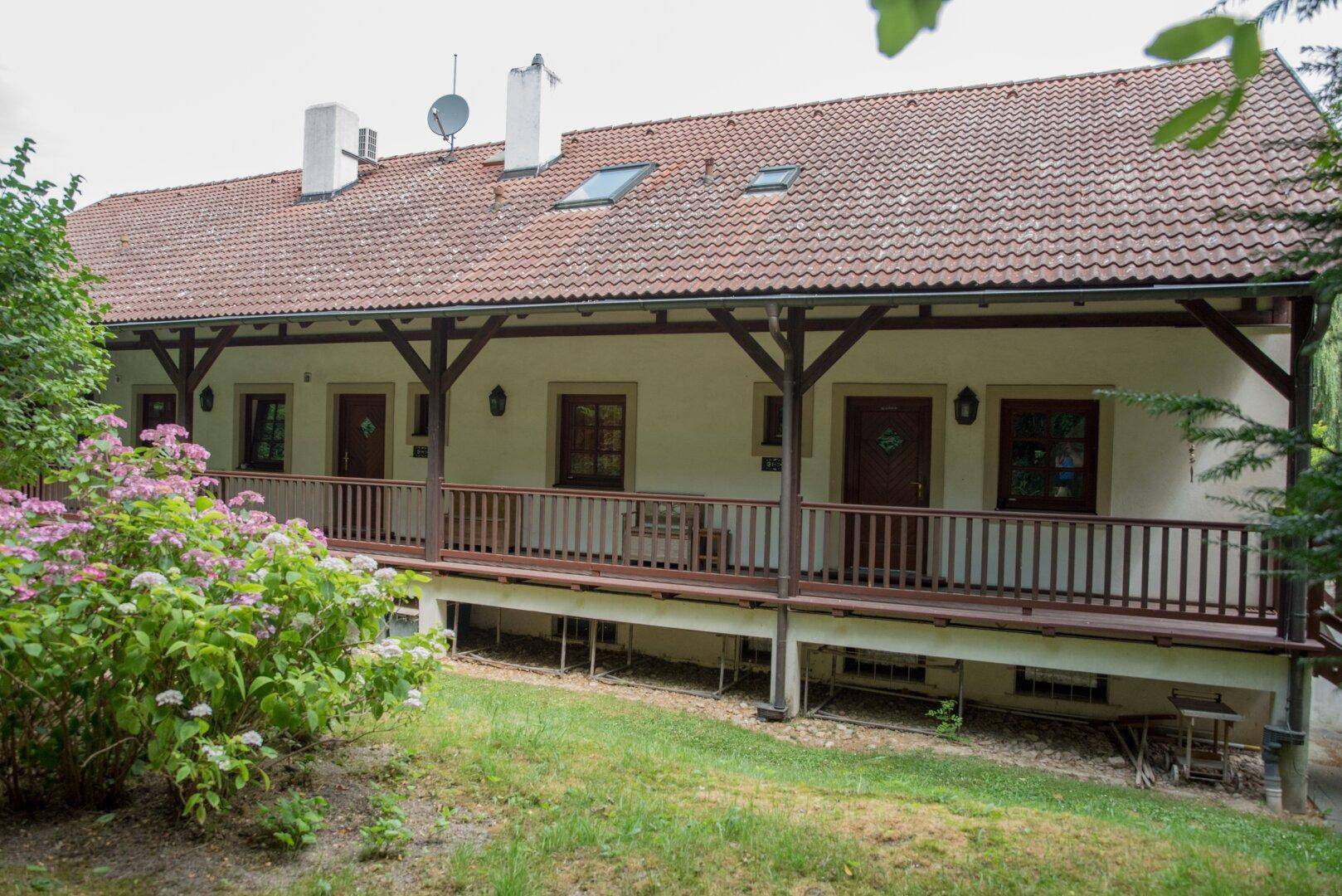 Lange, überdachte Loggia mit Holzbalken und Geländer, umgeben von blühenden Sträuchern.