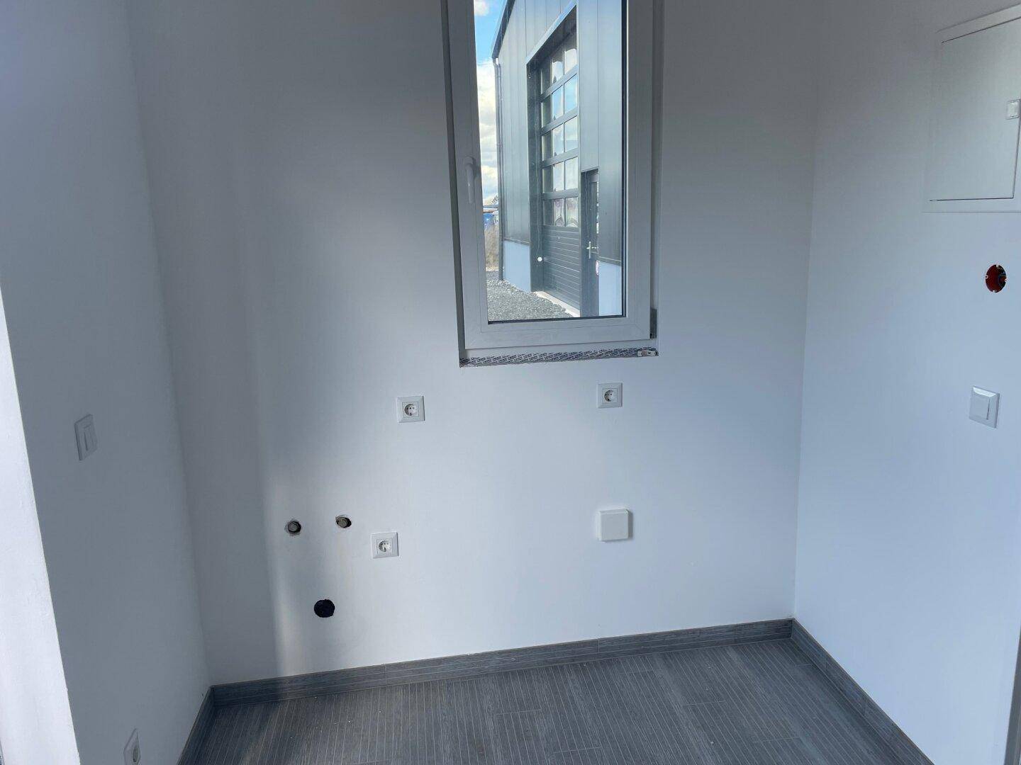 Leerer, kleiner Raum mit einem Fenster, das Tageslicht hereinlässt, und sichtbaren Elektroinstallationen.