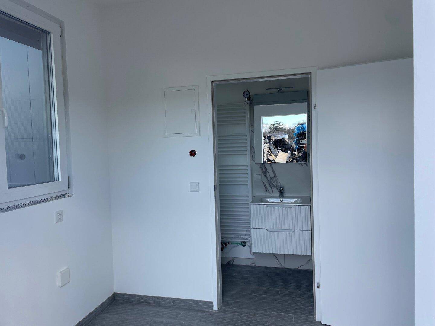 Kleiner, heller Raum mit Fenster und Blick in ein modernes Badezimmer mit Waschtisch und Spiegel.