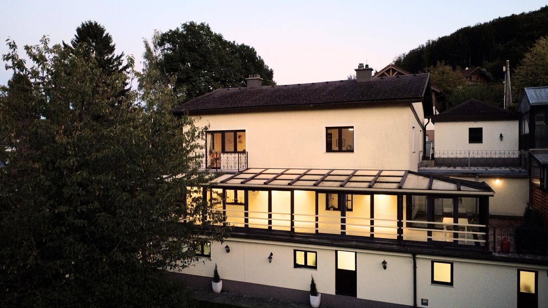Abendliche Ansicht des Hauses mit beleuchtetem Wintergarten und umgebender Natur.