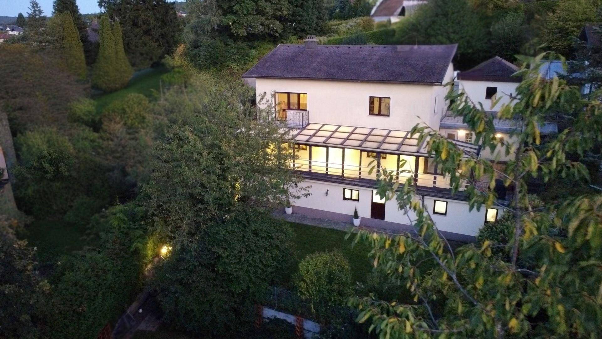 Ansicht des Hauses bei Dämmerung mit beleuchtetem Wintergarten und Gartenbereich.