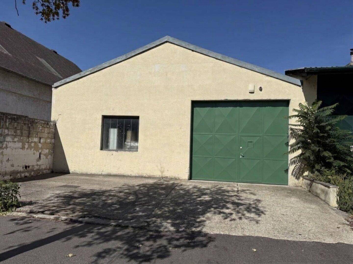Vorderansicht eines einfachen Lagergebäudes mit beiger Fassade, grünem Tor und einem kleinen Fenster.