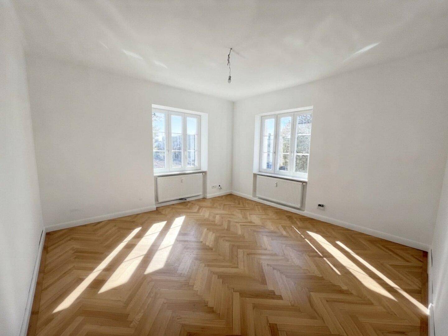 Helles Schlafzimmer mit Fischgrätparkett, weißen Wänden und zwei Fenstern für viel Tageslicht.