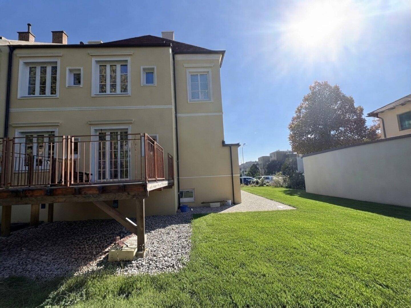Sonnige Rückseite des Gebäudes mit großer Rasenfläche und erhöhter Holzterrasse.