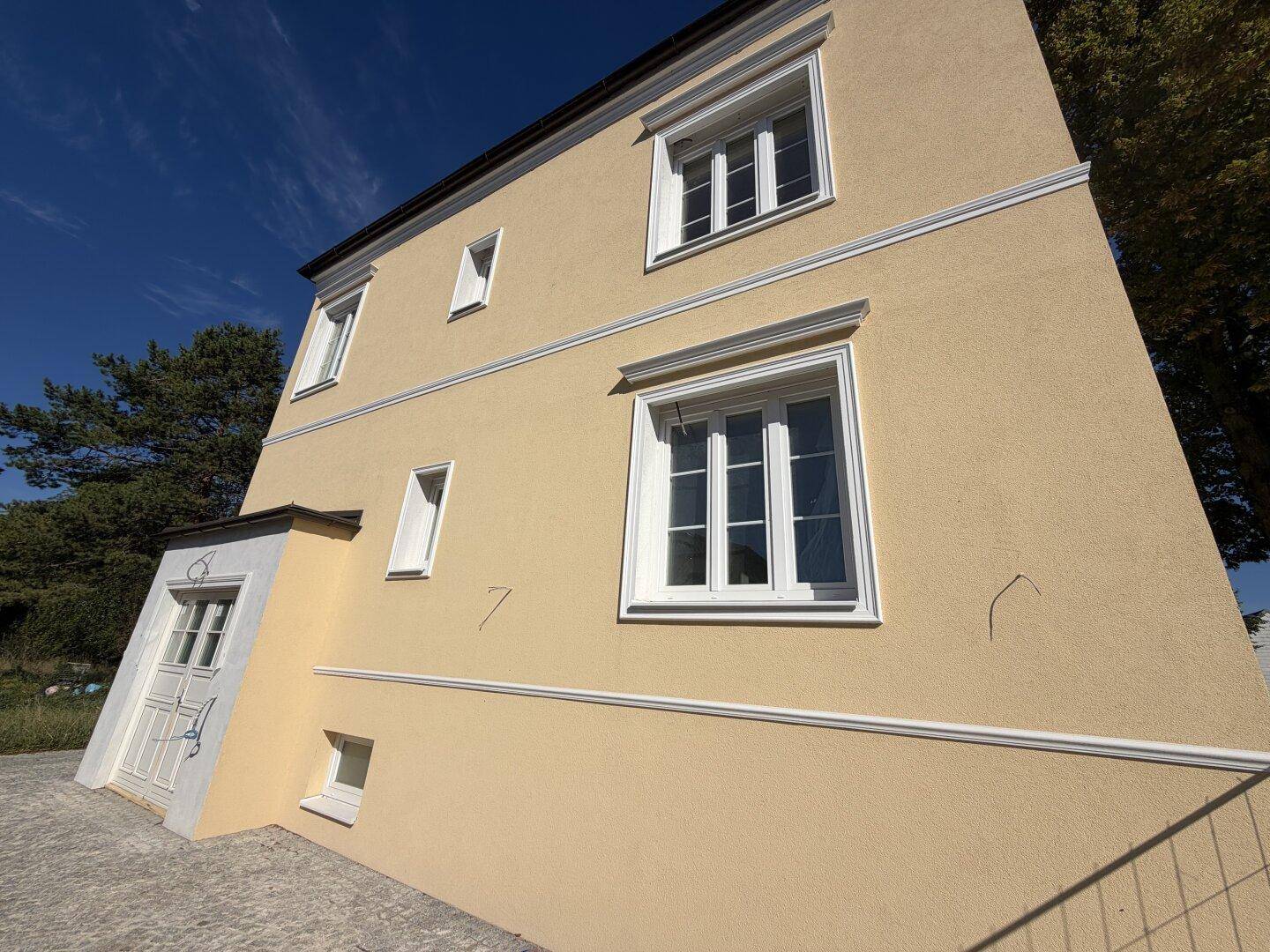 Seitenansicht des Gebäudes mit gelber Fassade, weißen Fenstern und einem Eingangsbereich.