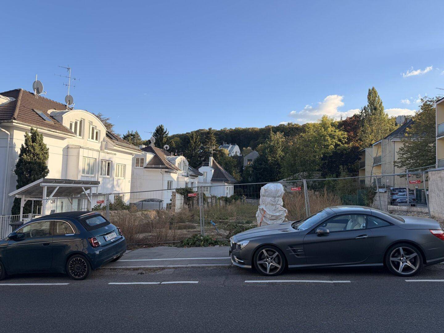 Panoramablick auf ein unbebautes Grundstück zwischen bestehenden Wohnhäusern mit hügeliger Landschaft im Hintergrund.