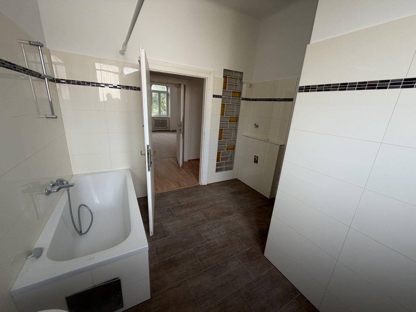 Badezimmer mit Badewanne, weißen Fliesen und dunklem Boden, bereit für eine Modernisierung.