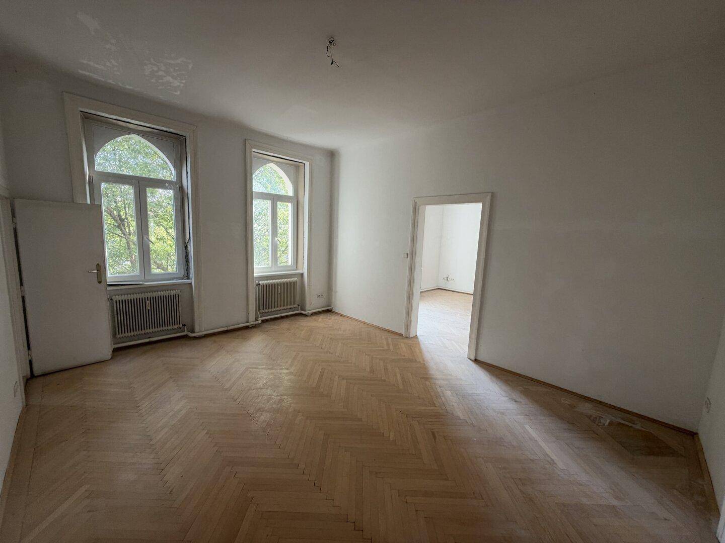 Geräumiges Zimmer mit Fischgrätparkett und zwei hohen Fenstern, die den Raum erhellen.