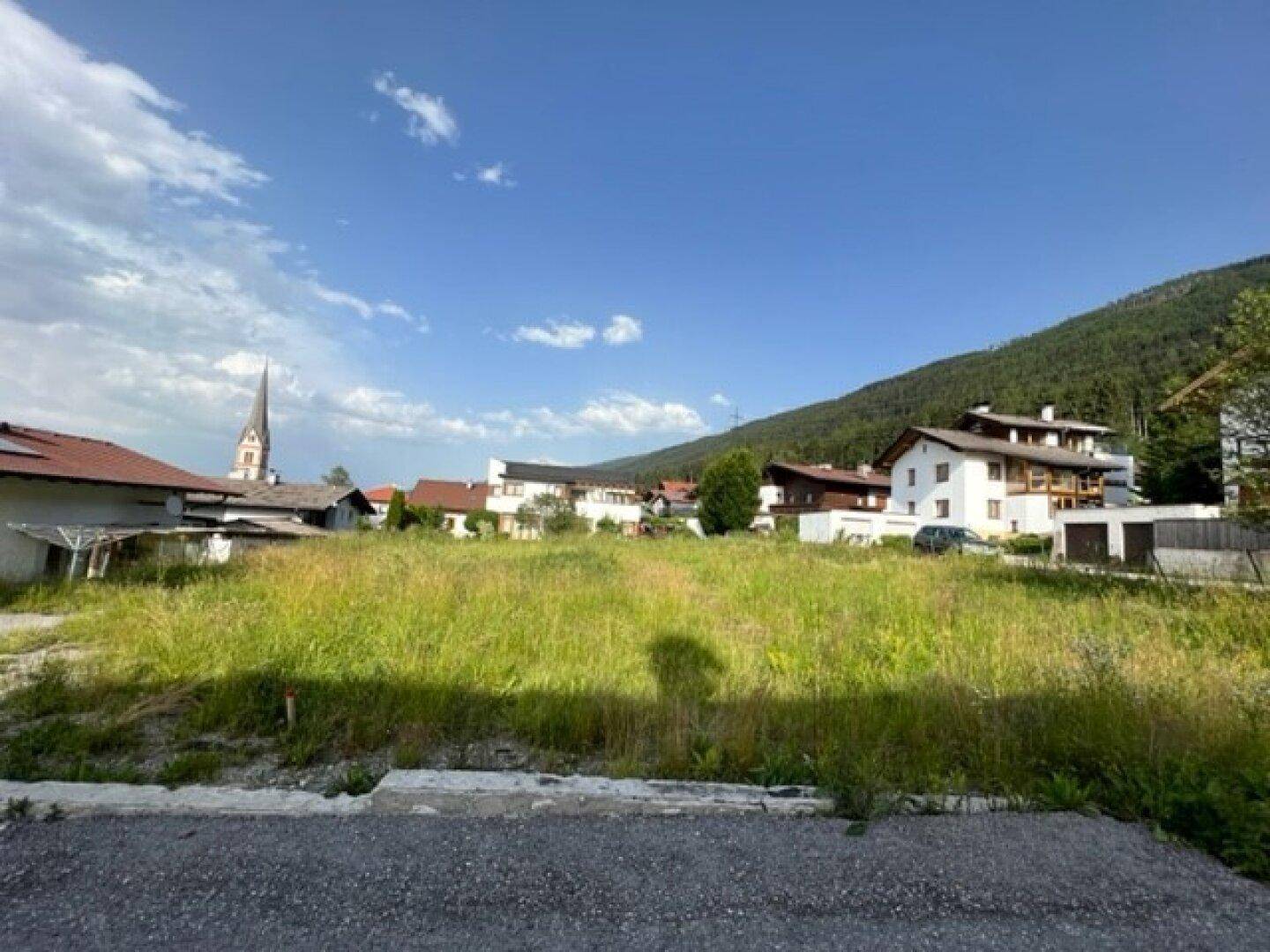 Grüne Wiese als Baugrundstück mit umliegenden Häusern und Kirchturm vor Bergkulisse.