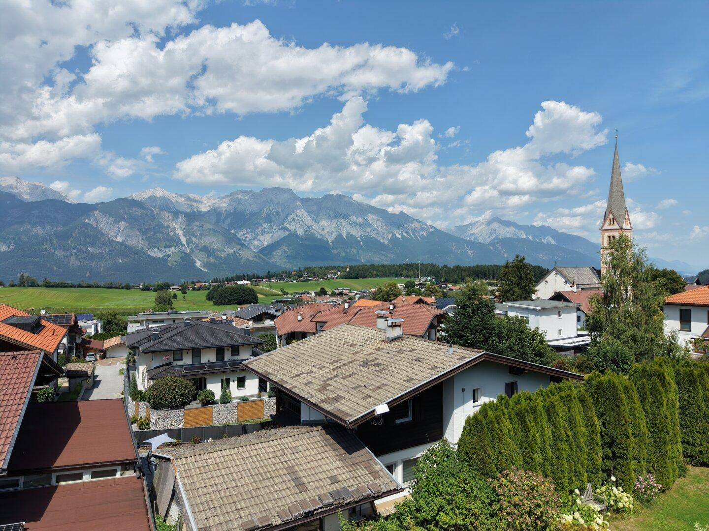 Panoramablick über eine sonnige Wohngegend mit Kirchturm und majestätischer Bergkette.