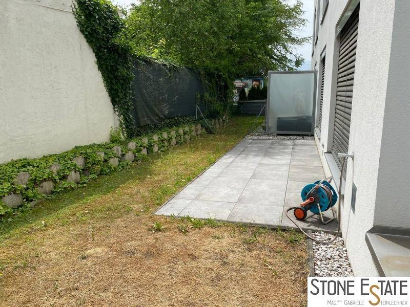 Seitlicher Gartenbereich mit gepflastertem Weg, Rasen und grüner Bepflanzung.