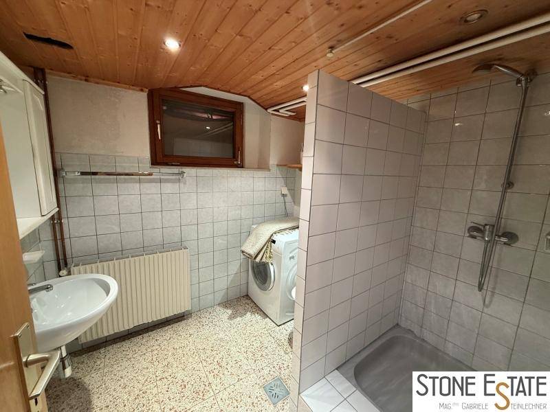 Badezimmer mit Dusche, Waschbecken, Waschmaschine und Fenster, teilweise gefliest.