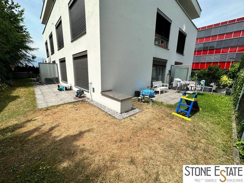 Gartenbereich mit Rasenfläche, gepflasterter Terrasse und Gebäudefassade.