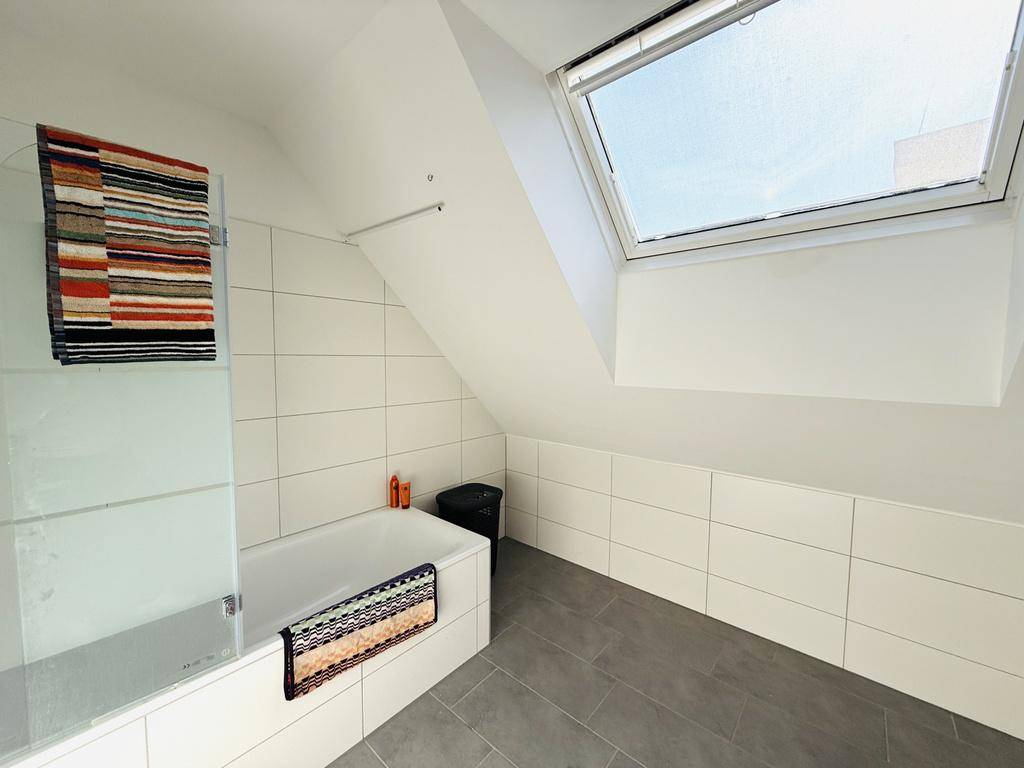 Badezimmer mit Badewanne, Duschabtrennung, Fliesenboden und einem Dachfenster in der Schräge.