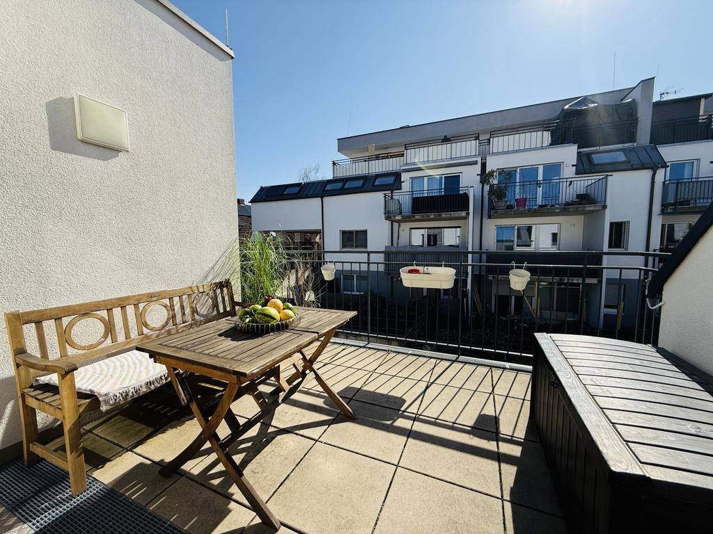 Sonnige Terrasse mit Holzbänken und Tisch, umgeben von anderen Wohngebäuden.
