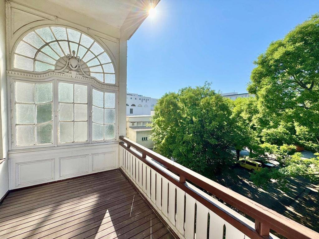Sonniger Balkon mit Holzdielen und historischem Fenster, umgeben von grünen Bäumen.