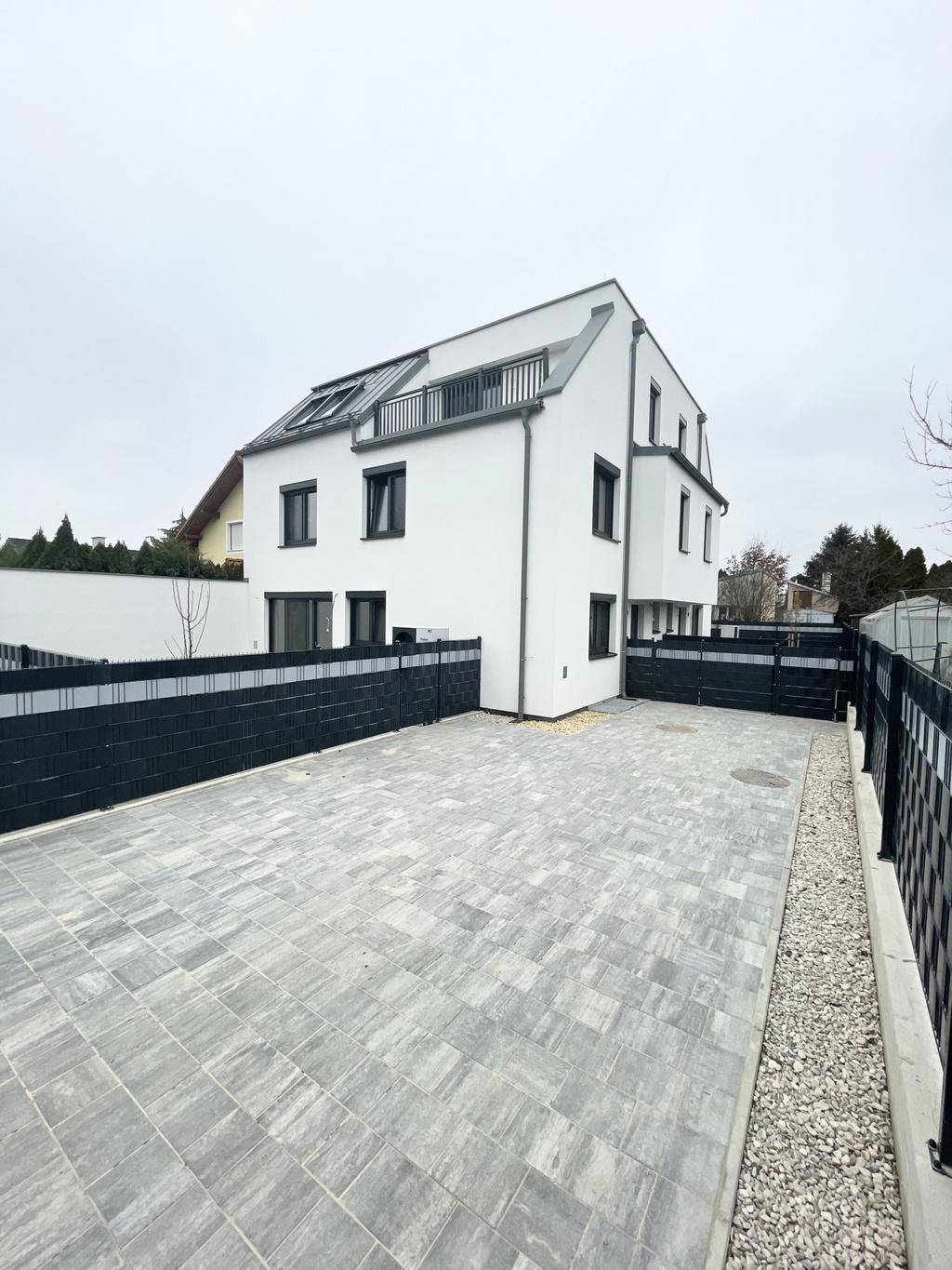 Seitenansicht eines modernen Hauses mit großzügiger gepflasterter Parkfläche und Zaun.