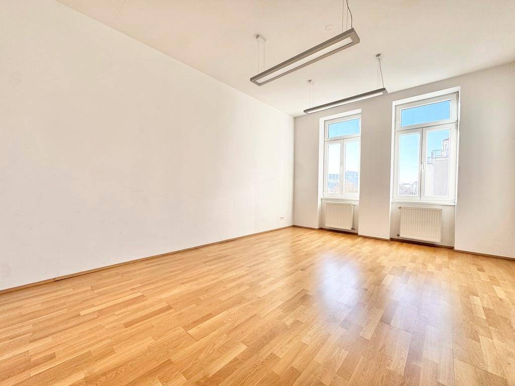 Geräumiger, heller Raum mit Holzboden, großen Fenstern und schlichten Deckenlampen.