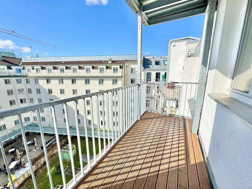 Langer Balkon mit Holzboden und Metallgeländer, bietet Blick auf umliegende Gebäude und einen Innenhof.